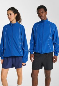 Blå sportjackor med vita pipingdetaljer, lättviktsmaterial, höga kragar och elastiska ärmslut, i kombination med mörka shorts.