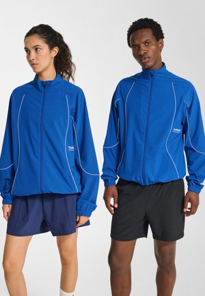 Blaue Sportjacken mit weißen Paspeldetails, leichter Stoff, hohe Kragen und elastische Bündchen, kombiniert mit dunklen Shorts.
