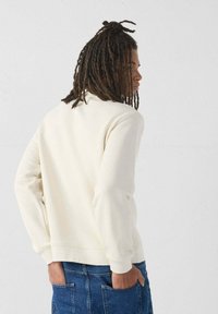 Sudadera color crema con un corte relajado, que cuenta con mangas largas, un escote redondeado y puños acanalados, combinada con jeans de mezclilla azul.