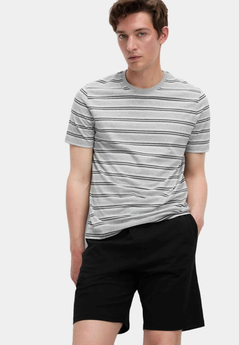 Mand iført en grå og sort stribet kortærmet skjorte og sorte shorts, stående med den ene hånd i lommen foran en ensfarvet lys baggrund.