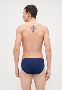 Marineblaue Schwimmhose mit glatter Textur, mit markiertem Branding an der Seite und figurbetontem Design. Verfügt über ein Fleur-de-Lis-Tattoo auf der Rückseite.