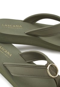 Olivgrüne Flip-Flops mit weichen Stoffriemen und einem metallischen Ringdetails. Das Fußbett ist glatt und mit einem geprägten Markenlogo in Gold versehen.