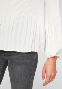 Witte geplooide blouse met lange, voluminieuze mouwen, vloeiende textuur en een ronde zoom, gecombineerd met grijze strakke jeans.