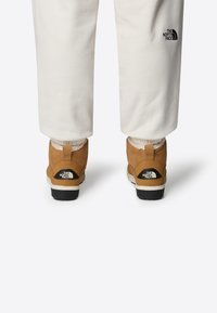 Brown boots with a textured suede finish, white faux fur lining, black rubber soles, and a prominent black logo on the heel. Cream pants.