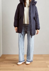 Giacca a vento blu navy con cappuccio, texture trapuntata, chiusura frontale con zip e tasche laterali, abbinata a jeans a gamba larga azzurri e sneakers bianche.