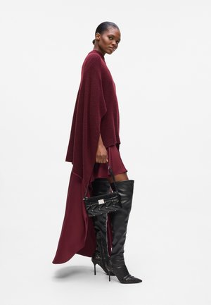 Burgundy knit poncho over a matching dress, paired with long black leather stiletto boots and a small black handbag with studs.