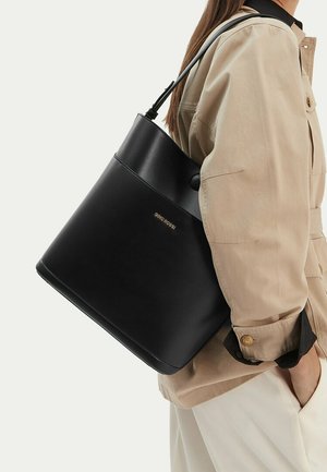 Mujer con chaqueta beige y pantalones blancos lleva un bolso grande tipo cubo negro con correa delgada sobre el hombro.