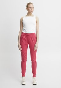 White ribbed tank top paired with high-waisted, pink tapered joggers featuring a drawstring. Model wears white sneakers. Simple background.
