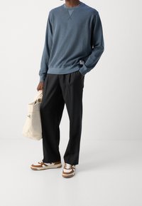 Blauwe sweatshirt van zacht materiaal met een ronde hals, gecombineerd met zwarte ruimvallende broek en bruin-witte sneakers. Draagt een crème tote bag.