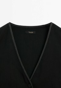 Schwarzes Kleidungsstück mit V-Ausschnitt, kontrastierenden weißen Nähten an den Kanten und einer einzelnen Knopfleiste. Innen ist ein Etikett sichtbar.