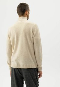 Uomo di spalle che indossa un maglione a collo alto beige e pantaloni grigio scuro su uno sfondo chiaro e semplice.