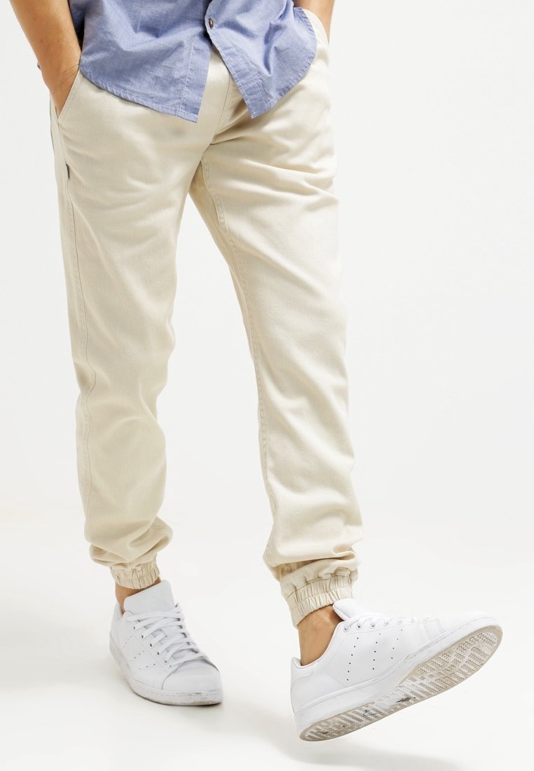 Homme portant un pantalon de jogging beige et des baskets blanches, levant le pied droit, les mains dans les poches, debout sur un fond blanc.
