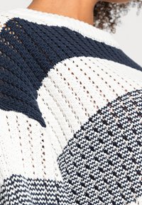 Gros plan sur une personne portant un pull en tricot blanc et bleu marine avec un motif ajouré et des blocs de couleurs géométriques.