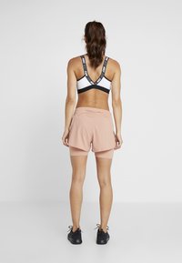 Czarny i biały sportowy biustonosz z paskami z logo, w połączeniu z różowymi szortami sportowymi z bocznymi kieszeniami. Modelka stoi tyłem do kamery.