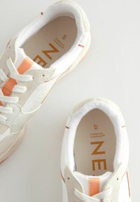 Białe i beżowe sportowe sneakersy z pomarańczowymi akcentami, wykonane z materiałów skórzanych i tekstylnych, okrągły czubek, gruba podeszwa, sznurowany fason oraz miękka wkładka.