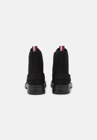 Botas de ante negras hasta el tobillo con una suela negra gruesa. Presentan un tirador en la parte posterior de rayas rojas, blancas y azules en contraste y un detalle de correa.