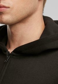 Sudadera negra con cremallera en la parte delantera, con una textura de tela suave y un cuello relajado. Vista de cerca del área del cuello y la barbilla.
