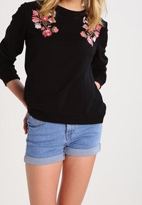 Sudadera negra con bordado floral en rosa y blanco en el pecho, combinada con pantalones cortos de mezclilla con dobladillos enrollados.