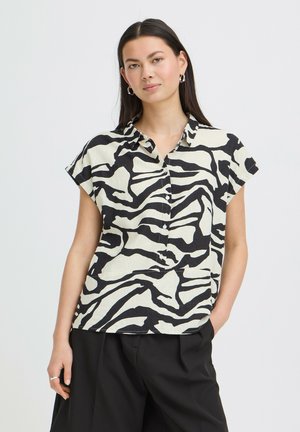 Young woman with long dark hair wearing a short-sleeve black and white zebra print button-up shirt and black pants, posing with hand in pocket.