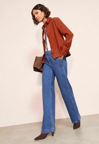 Rozsdabarna gombos ing, fehér felső, kék bőszárú farmer, barna kézitáska és barna hegyes orrú magassarkú, semleges háttér előtt.