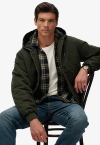 Zielona bombka z kapturem, z czarnym wnętrzem, noszona na niebieskiej i żółtej koszuli w kratę, białym t-shircie i niebieskich dżinsach.