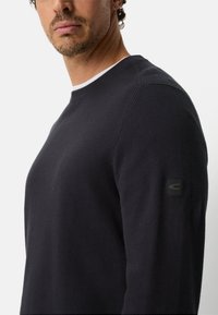 Pull noir texturé avec un col rond, un col intérieur blanc contrastant et un logo accent noir sur la manche gauche.