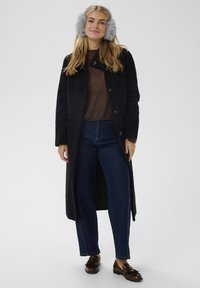 Lungo cappotto nero con tessuto strutturato, maglione marrone, jeans blu a gamba larga e mocassini marroni. Paraorecchie grigi in pelliccia aggiungono calore.