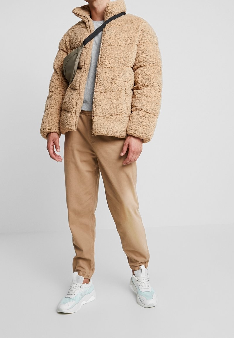 Beige Fleecejacke mit hohem Kragen, kombiniert mit einem hellgrauen Sweatshirt und sandfarbenen, zulaufenden Hosen. Weiße und blaue Sneaker vervollständigen das Outfit.
