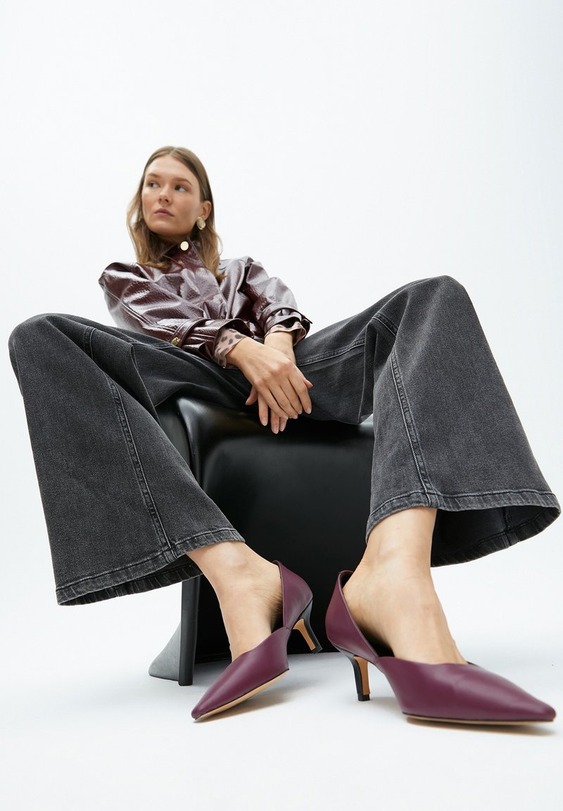 Donna seduta su una sedia nera, indossa una giacca di pelle viola, jeans neri larghi e tacchi a spillo viola, guarda di lato su uno sfondo bianco.