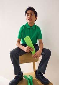 Žalia polo marškinėliai su trimis sagomis, pilkos kelnės ir juodi sportbačiai. Laiko šviesiai žalius popieriaus juosteles, sėdi ant medinės kėdės.