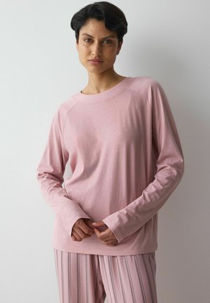 Person mit kurzen dunklen Haaren, die ein hellrosa langärmeliges Oberteil und rosa gestreifte Hosen trägt, steht vor einem schlichten hellen Hintergrund.
