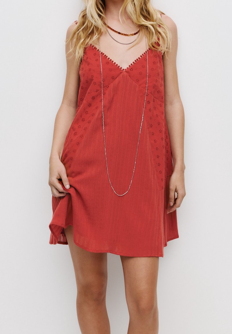 Femme portant une robe rouge sans manches avec des découpes florales et des colliers superposés, tenant l'ourlet de la robe d'une main.