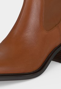 Gros plan sur l'avant et le côté d'une bottine en cuir beige avec une semelle noire et un petit talon empilé.