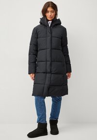 Zwarte pufferjas met een capuchon, voorzien van een doorgestikte ontwerp, drukknoopsluiting en zijzakken. Gecombineerd met blauwe jeans en zwarte laarzen.