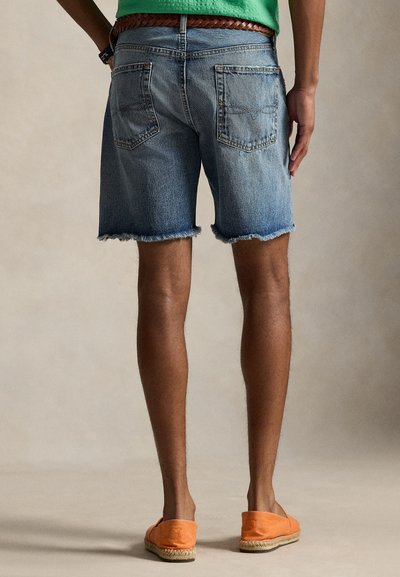 Moški v raztrganih džins kratkih hlačah z razpokanimi robovi, zeleni srajci, pletenem rjavem pasu in oranžnih espadrilih stoji ob nevtralnem ozadju, pogled od zadaj.