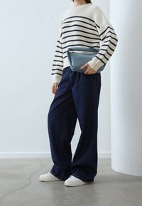 Pull rayé crème et marine associé à un pantalon en denim foncé à jambes larges. Le mannequin tient une pochette en cuir bleu doux. Des baskets blanches complètent la tenue.