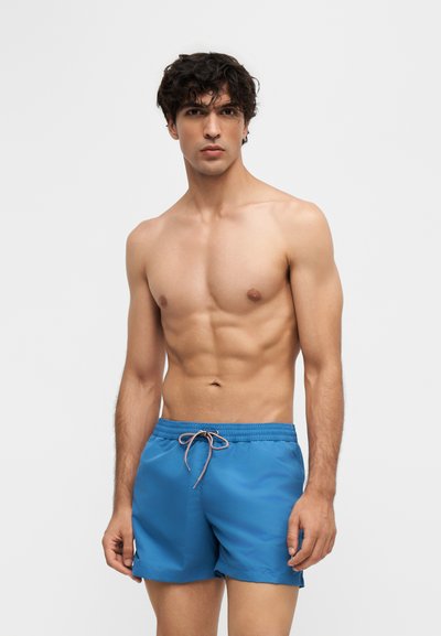 Jeune homme aux cheveux bouclés foncés portant un short de bain bleu, debout torse nu devant un fond clair uni.