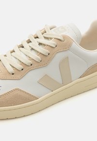 Weiße Leder-Sneaker mit beigen Wildlederspitzen, runder Kappe und flachen Schnürsenkeln. Merkmale ein auffälliges beiges Logo an der Seite und eine strukturierte Sohle.