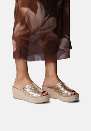 Noge, obute v sijoče zlato tkaninske natikače s platformo in bež podplati iz juta, pod rjavim cvetličnim prosojnim krilom.