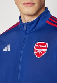Blaue Sportjacke mit roten Schulterstreifen, Reißverschluss und einem gestickten roten und weißen Arsenal-Logo auf der Brust.