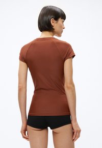 Rövid sötét hajú nő rozsdabarna, testhezálló rövid ujjú felsőben és fekete rövidnadrágban, háttal állva fehér háttér előtt.