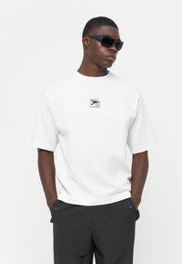 Biely bavlnený tričko s krátkymi rukávmi, s malým čiernym potlačeným logom na hrudi. Model nosí tmavé slnečné okuliare a čierne nohavice.