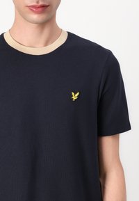 Tricou cu mânecă scurtă de culoare bleumarin, cu gât rotund de culoare bej. Prezintă un mic logo brodat cu o pasăre galbenă pe pieptul stâng. Material din bumbac.