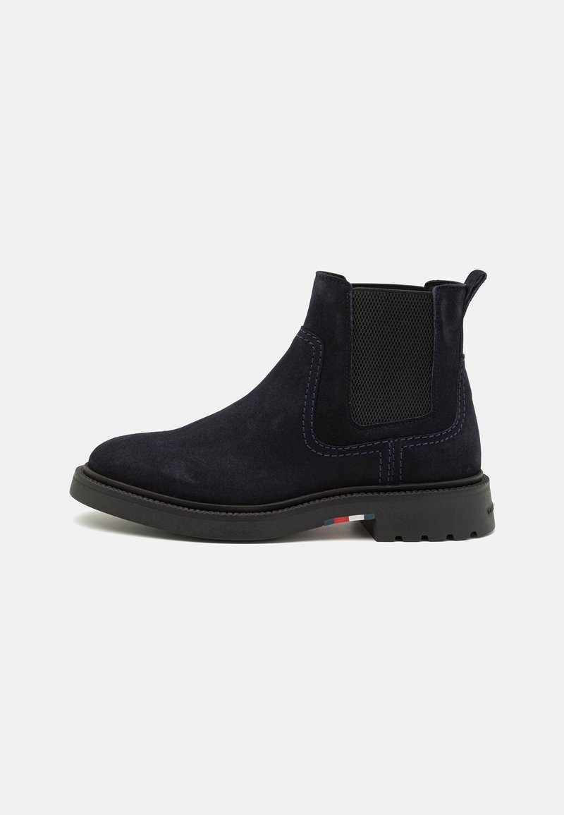 Bottine Chelsea en daim noir avec panneau latéral élastique, languette, petit talon et semelle en caoutchouc avec un petit détail rouge, blanc et bleu.
