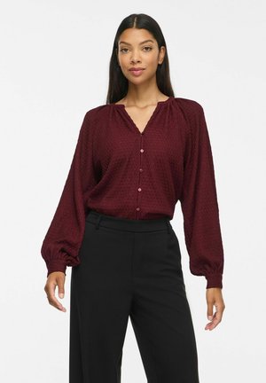 Femme portant un chemisier boutonnière bordeaux texturé avec de longues manches bouffantes et un pantalon noir taille haute, debout devant un fond blanc.