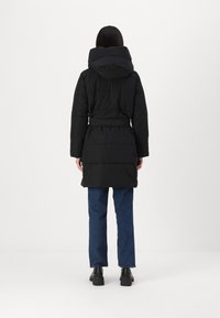 Abrigo de plumas negro con cuello alto, cintura ceñida y textura acolchada. Lleva puesto unos vaqueros azules y botas negras gruesas.