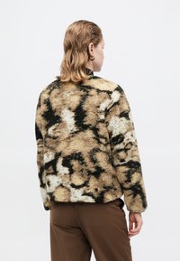 Chaqueta de piel sintética con un patrón abstracto en beige y negro, cuello redondo y mangas largas, combinada con pantalones marrones. Textura suave evidente.