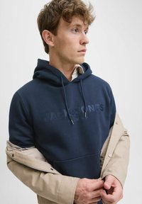 Sudadera azul marino con capucha y bolsillo frontal, que presenta un logo en relieve. Capa con una chaqueta exterior beige clara con puños a presión.