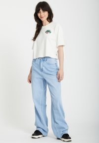 T-shirt blanc court avec broderie florale, assorti à un jean large bleu clair. Le modèle porte des baskets noires. Fond blanc uni.