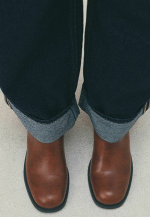 Person wearing cuffed dark jeans and brown leather square-toe boots standing on a light floor.